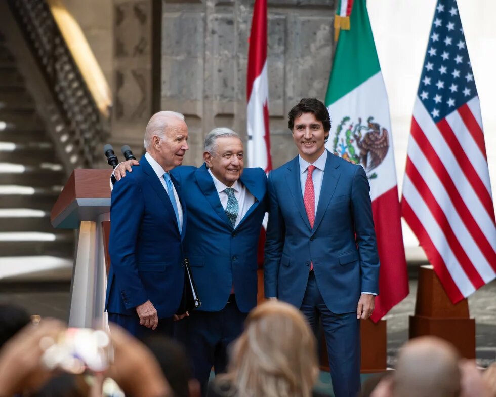 Foto: Joe Biden, Andrés Manuel López Obrador und Justin Trudeau stehen nebeneinander, lächeln und umarmen sich vor Flaggen der USA, Mexiko und Kanada.