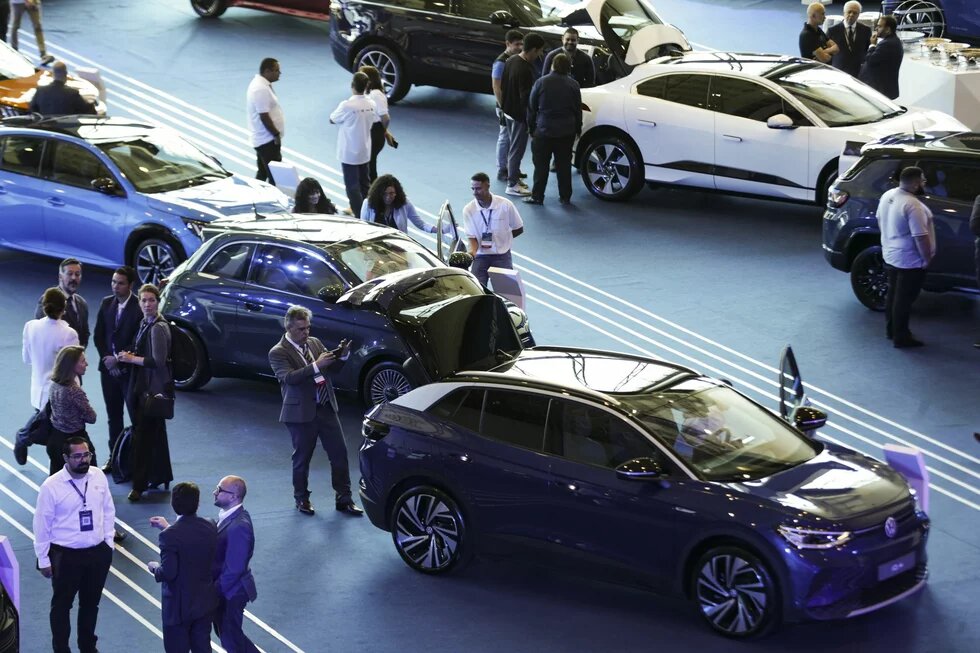 Foto: Menschen betrachten Elektroautos in einer Ausstellungshalle. Fahrzeuge sind geöffnet, einige Personen diskutieren oder machen Fotos.