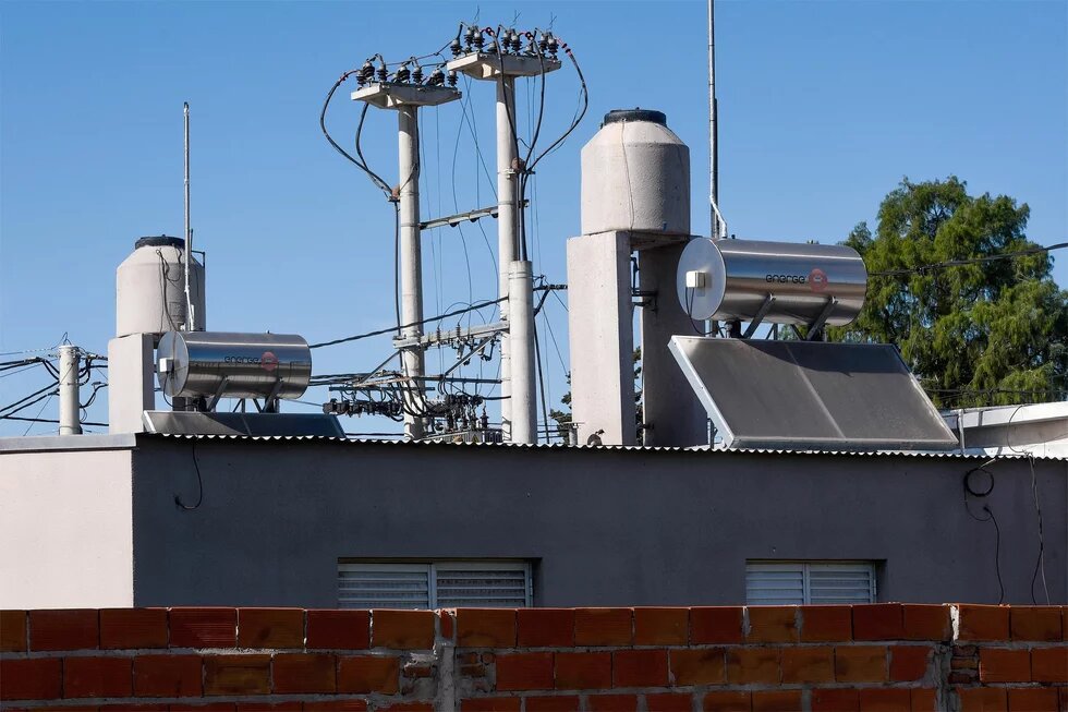 Foto: Flachdach mit Wassertanks, Solarpanelen und Strommasten im Hintergrund, teilweise von einer roten Ziegelmauer verdeckt. Klarer Himmel